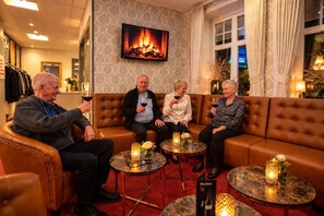 A group of people seated on a brown leather couch in a warmly lit room with a fireplace, a television mounted on the wall, and a small table with candles and a vase of flowers. - Hotel Kløver Es (Rodekro)