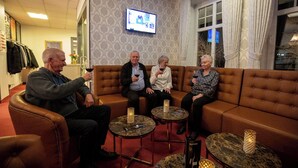 A group of people seated on a brown leather sofa in a room with patterned wallpaper, a television mounted on the wall, and a window with curtains. - Hotel Kløver Es (Rodekro)