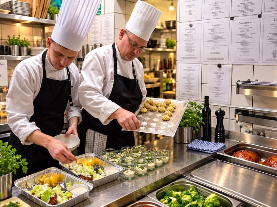 Chefs preparing food in a kitchen with stainless steel counters and menu boards on the wall.