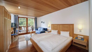 Een slaapkamer met een groot bed, een houten hoofdbord, een houten garderobe, een kleine tafel met een stoel, een bank en een balkon met uitzicht op bomen.