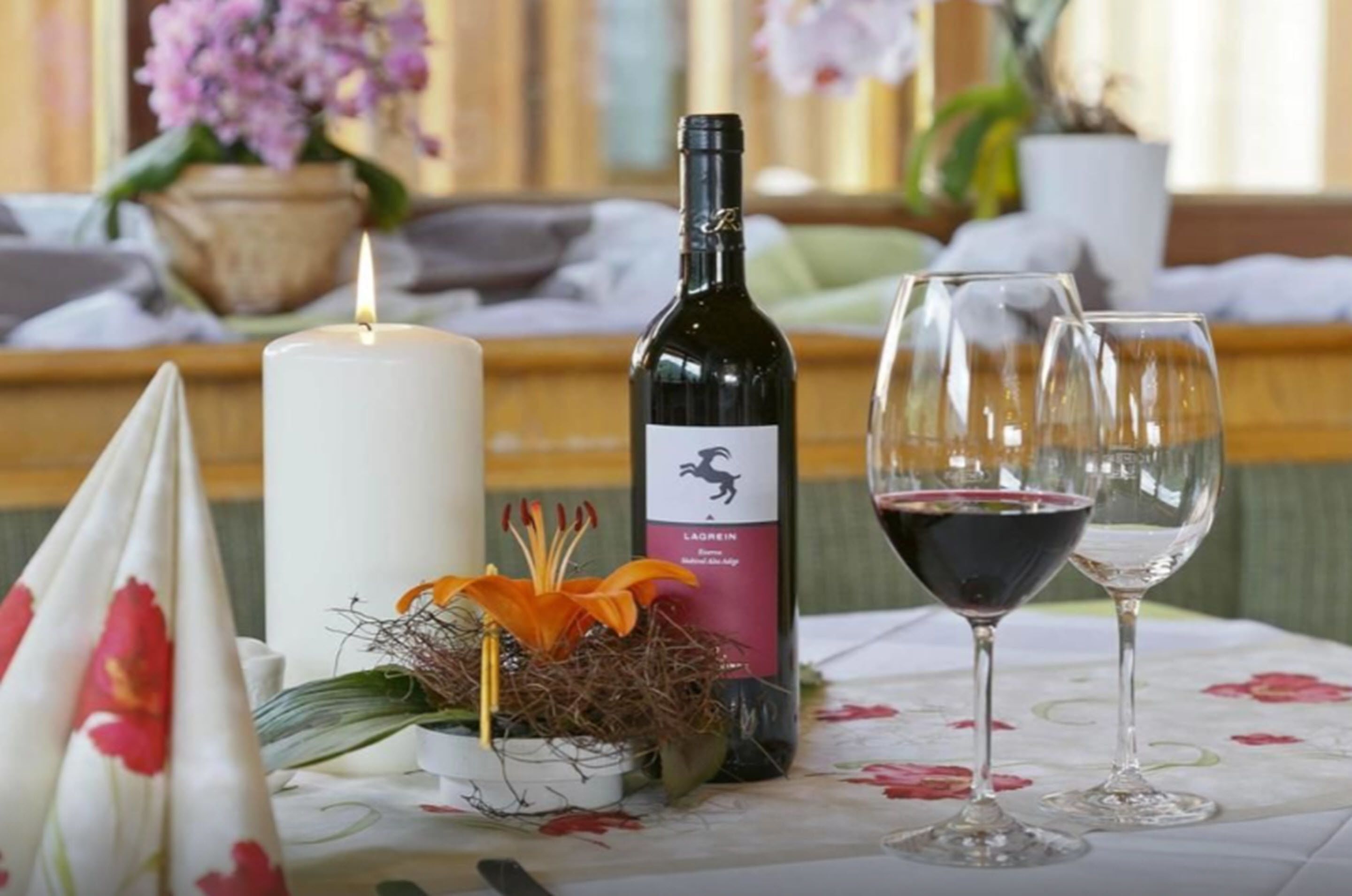 Una mesa con una botella de vino, dos copas, una vela encendida y un arreglo floral.