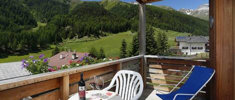 Una terraza de madera con una mesa y sillas, con vistas a un paisaje montañoso donde se distingue una casa al fondo.