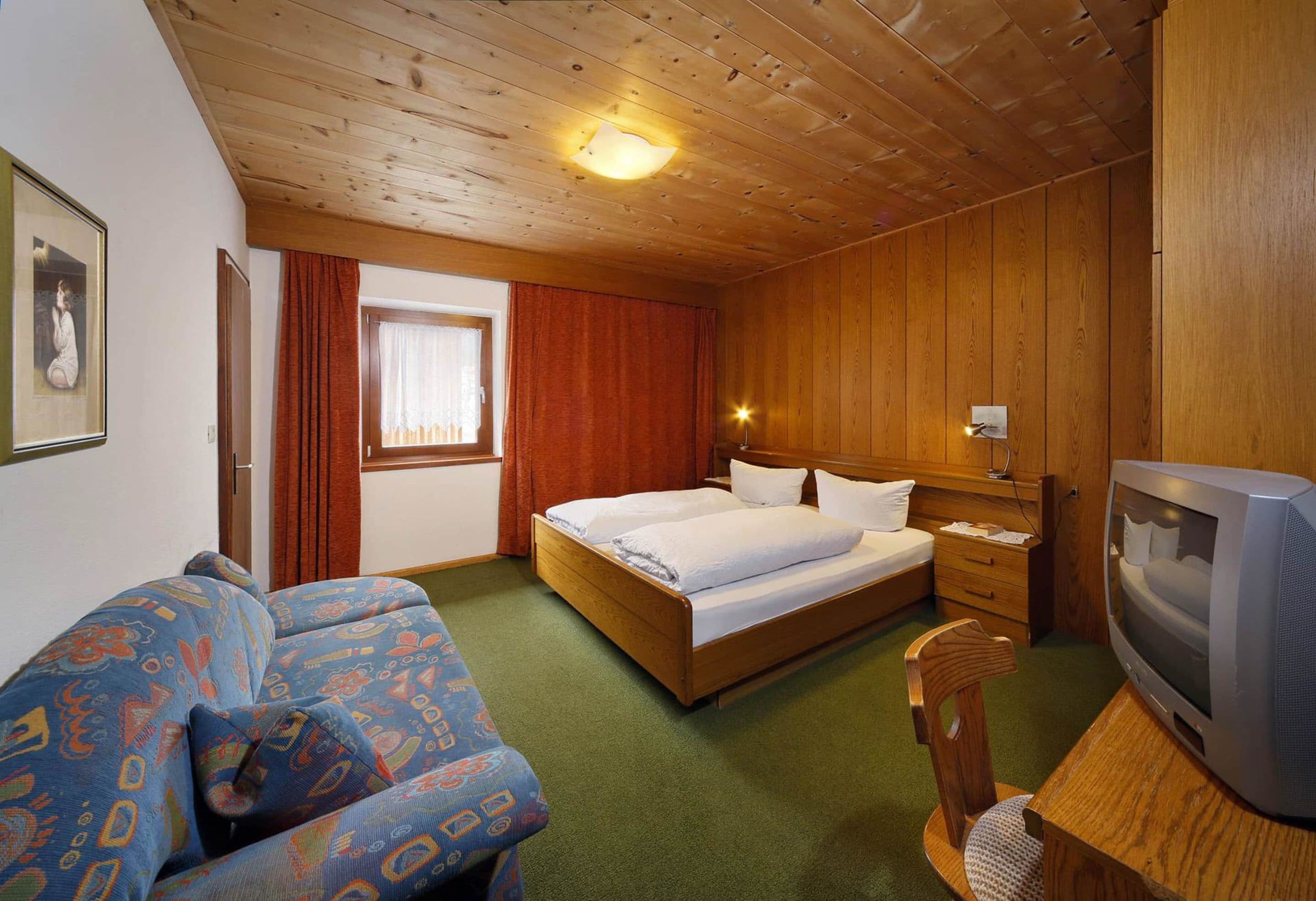 A room with a wooden ceiling, a bed with white linens, a television, and a patterned sofa.