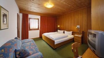 Quarto com teto de madeira, cama com lençóis brancos, televisão e sofá estampado.