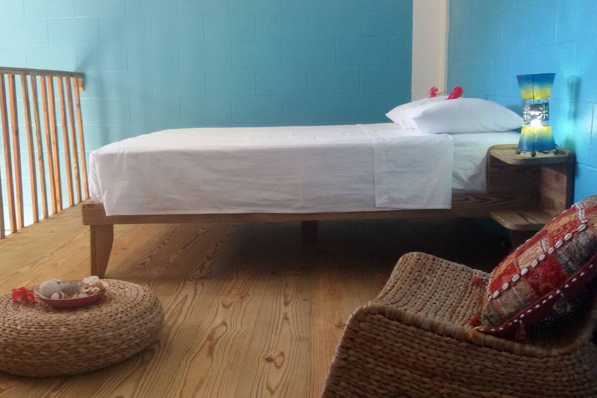 Una habitación con cama, piso de madera, un otoman tejido y una mesita de noche con lámpara.