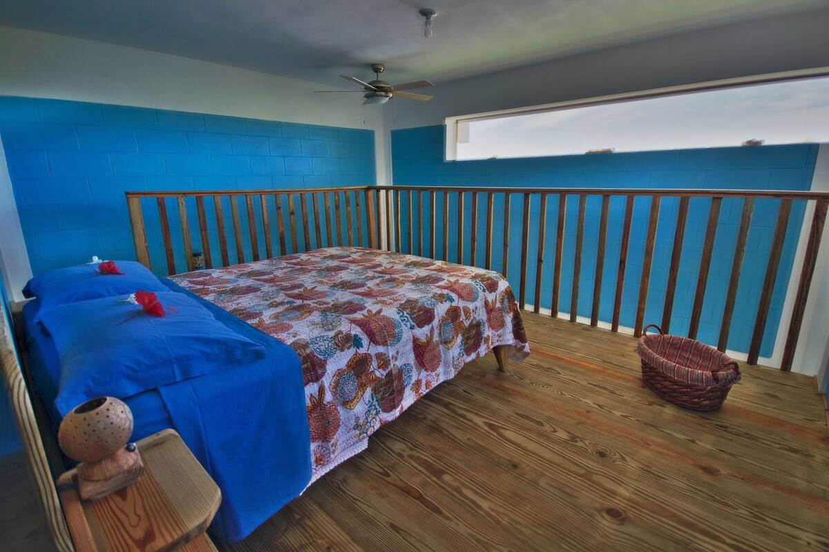 Un dormitorio con pared azul, estructura de cama de madera, colcha estampada y ventilador de techo.