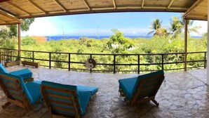 A covered outdoor area with stone flooring, two wooden lounge chairs with blue cushions, and a view of the ocean and greenery. - B&B El Mirador al Mar (Sosúa)
