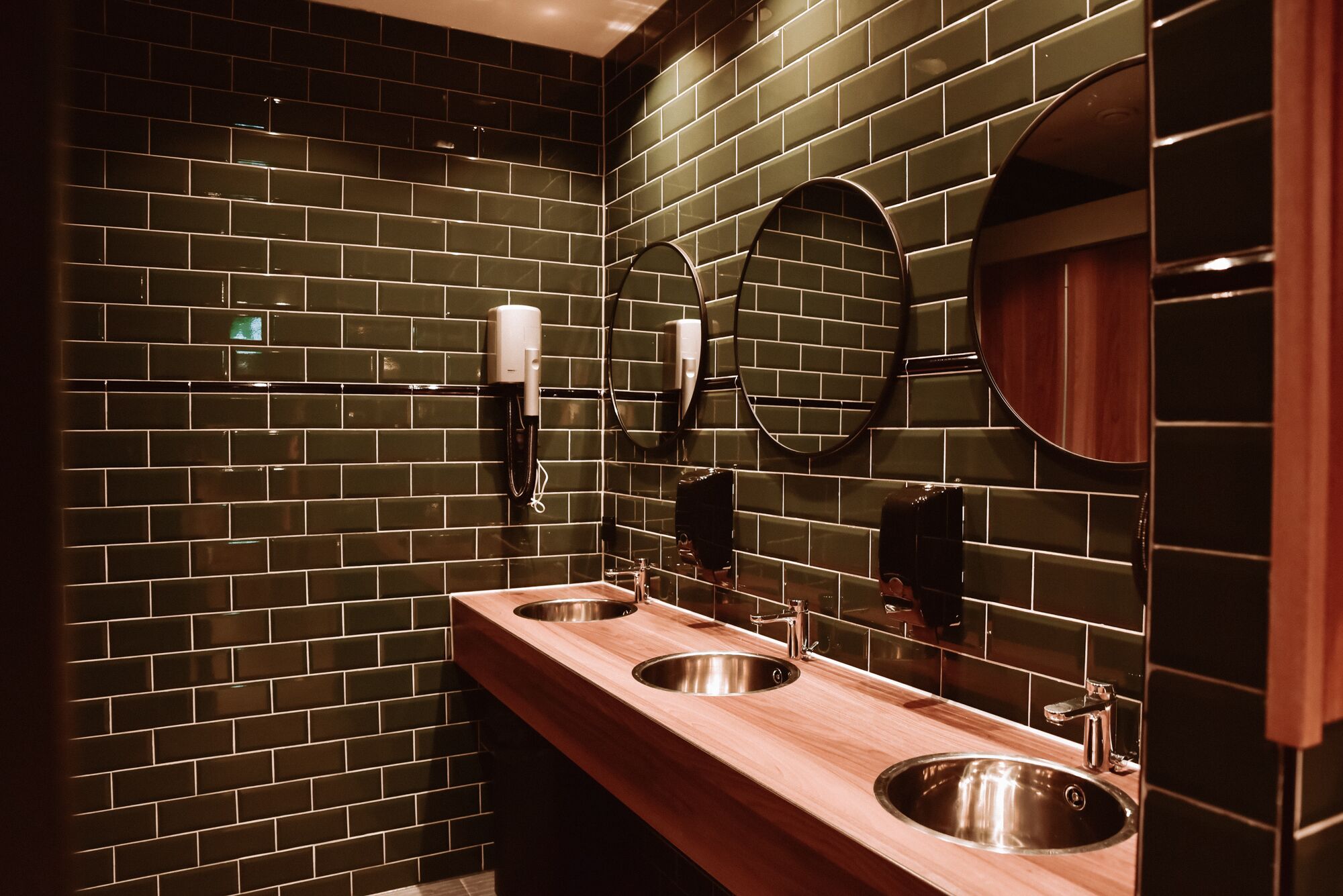 A modern bathroom with dark green tiled walls, two sinks with stainless steel basins, and round mirrors.