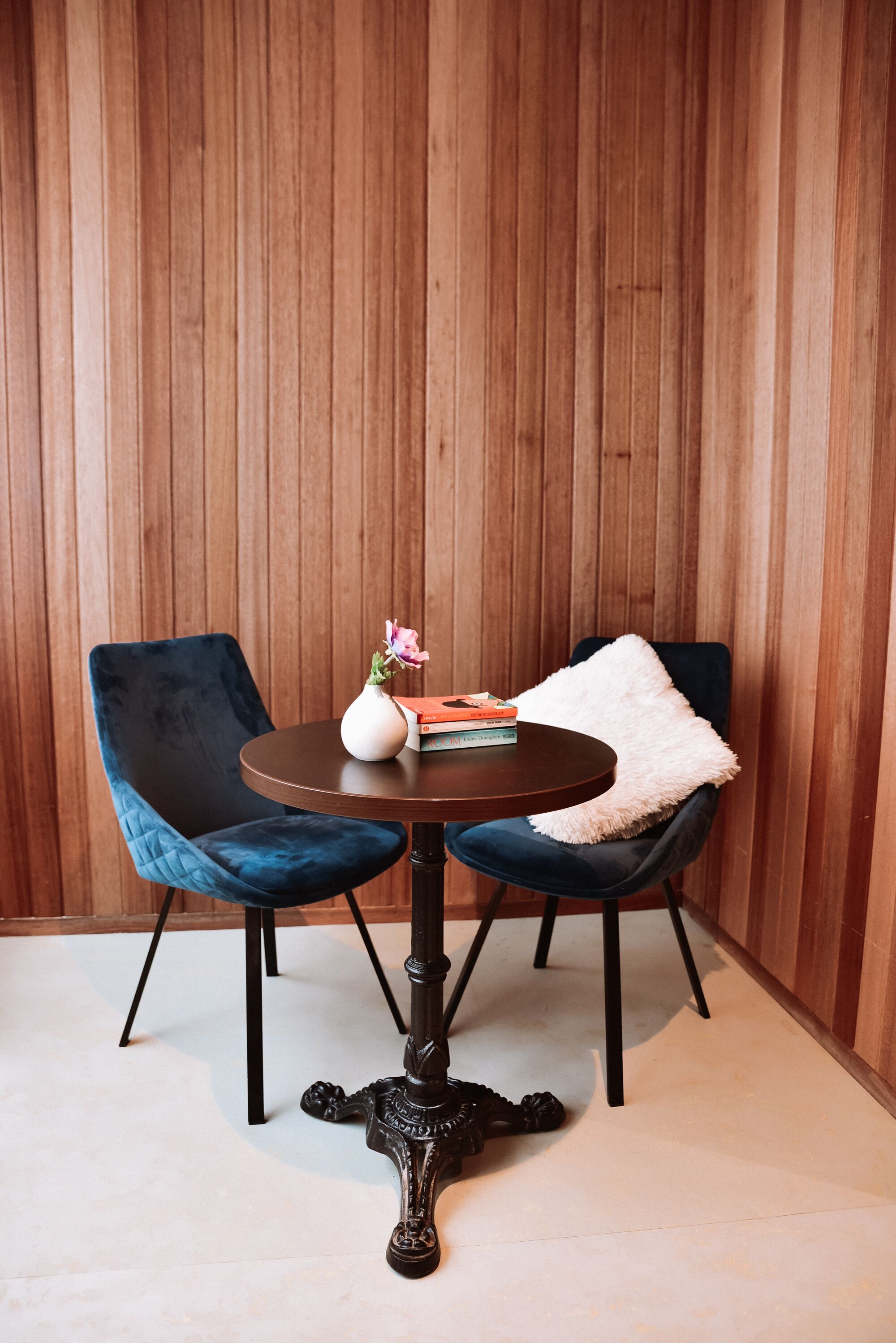 A room with a round table, two chairs, a vase with flowers, and a stack of books.