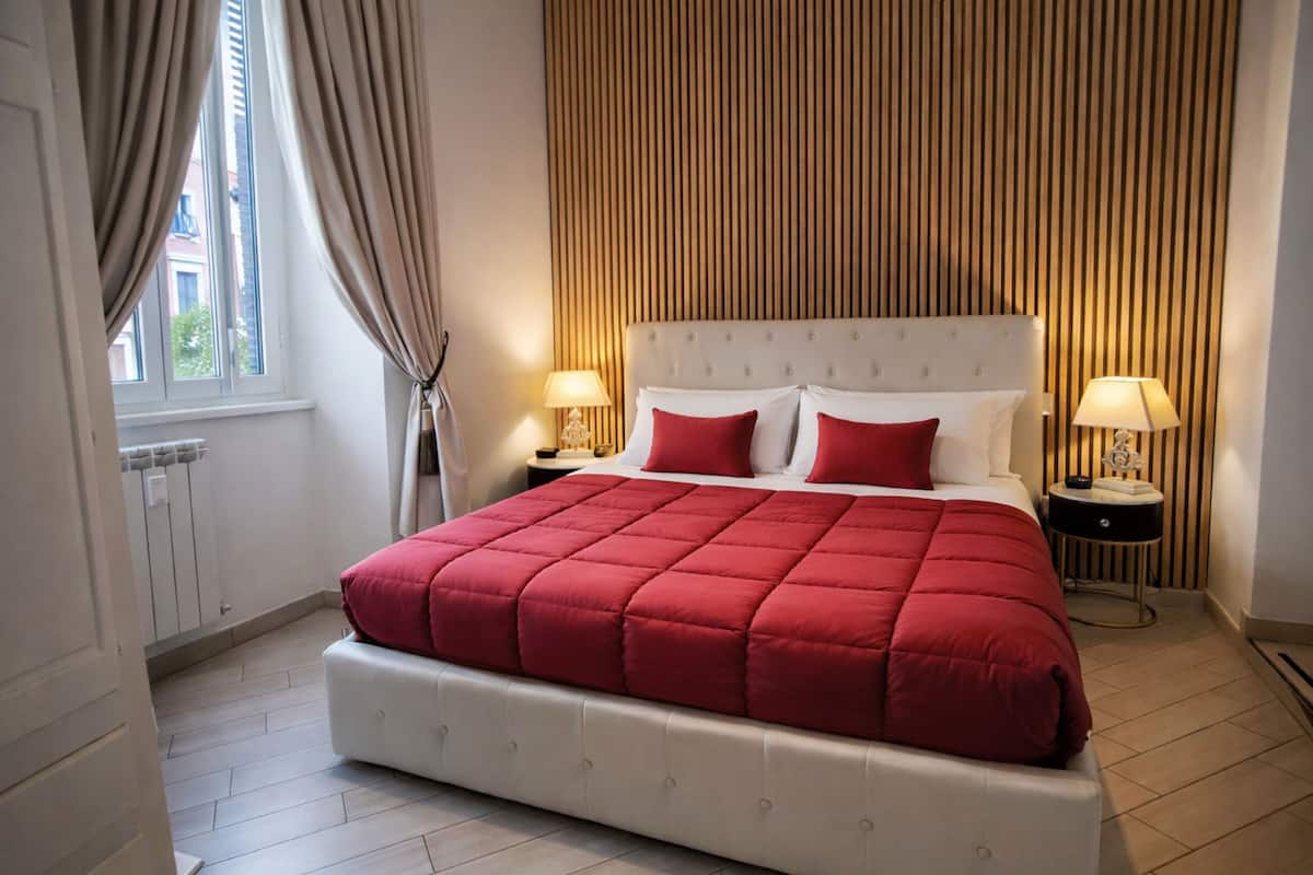 A hotel room with a large bed, two bedside tables with lamps, a window with curtains, and a wooden panel wall.