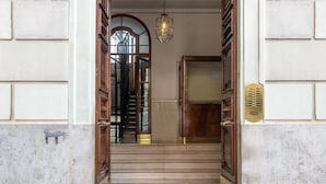 A marble staircase leading to a wooden door with a brass plate, a hanging light fixture, and a view of a staircase and a window in the background. - Rome Central Rooms Guest House (Rome)