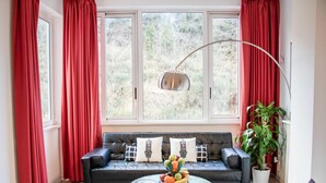 A living room with a black leather sofa, a glass coffee table with fruit, red curtains, and a view of trees through large windows. - Gallery (Rome)