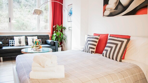A hotel room with a bed, a sofa, a round coffee table, and a large window with curtains. - Gallery (Rome)