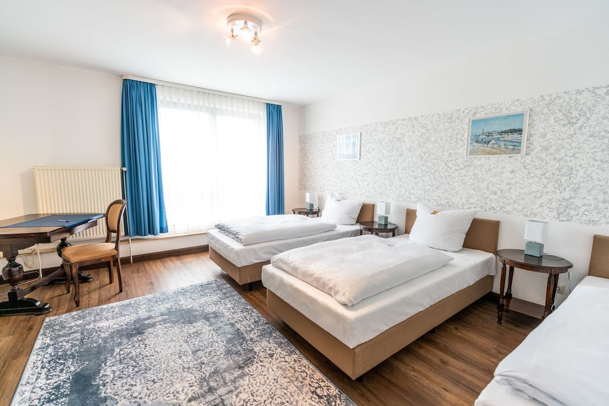 Une chambre d’hôtel avec deux lits, un bureau, une chaise, un tapis, un tableau et une fenêtre avec des rideaux bleus.