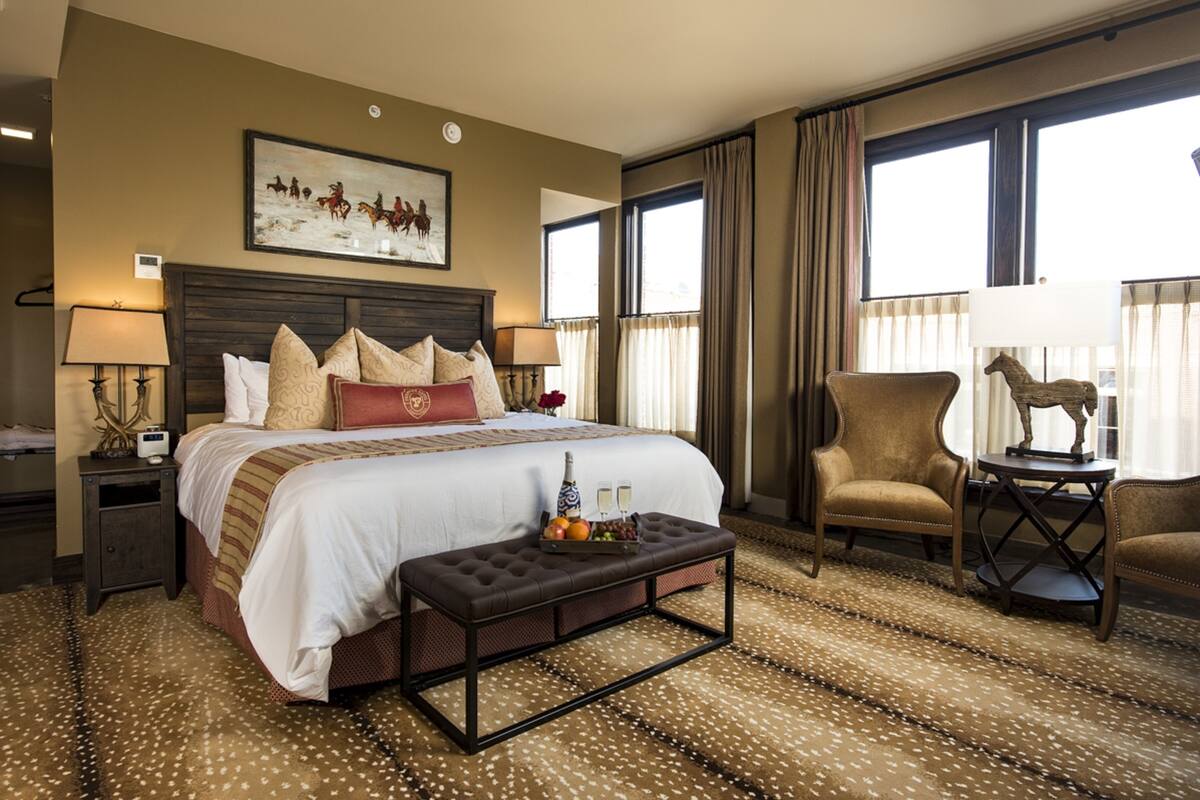 A hotel room with a large bed, a wooden headboard, a bench, a chair, a small table with a statue, and a painting on the wall.