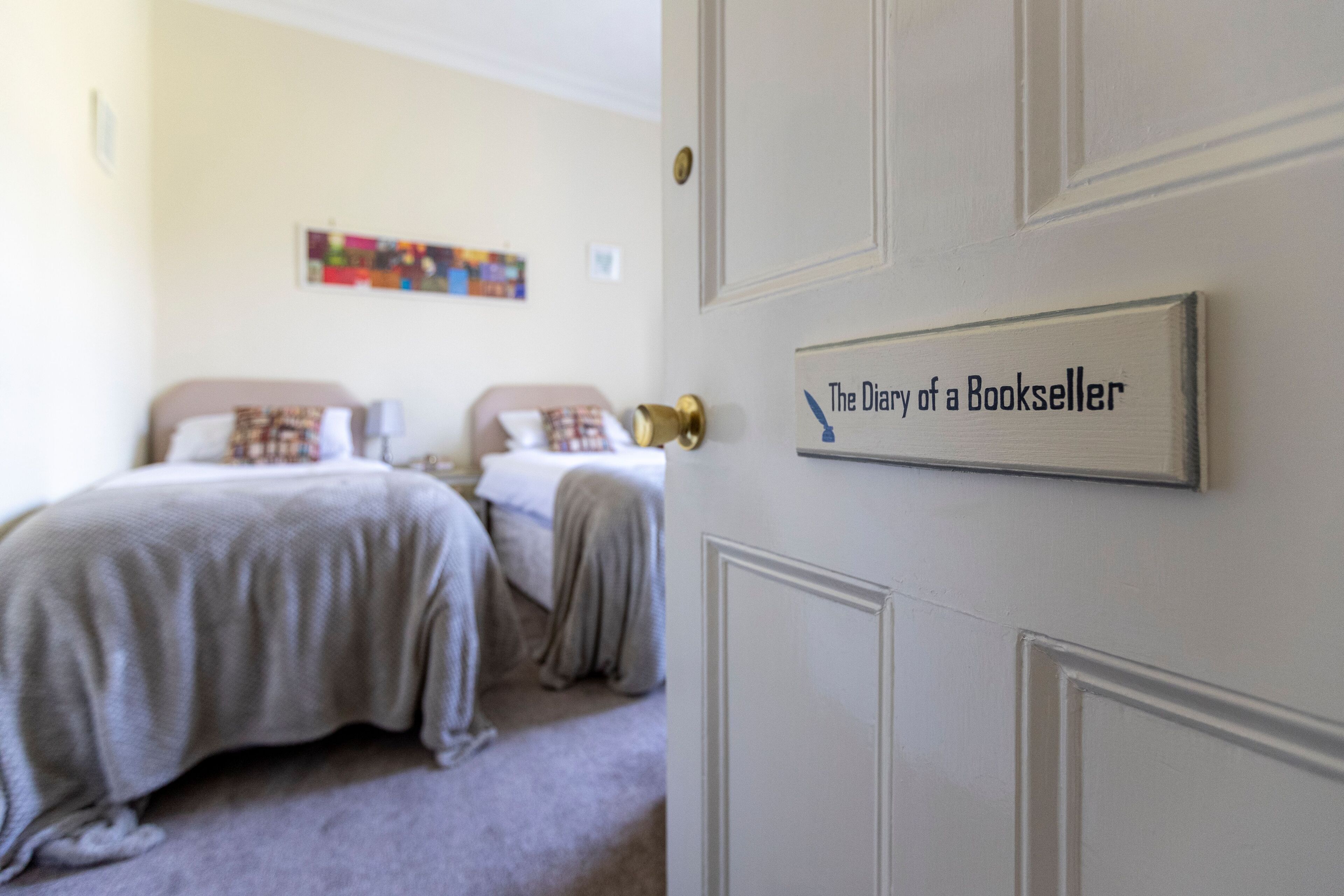 A hotel room with two beds, a door with a sign reading "The Diary of a Bookseller," and a colorful artwork on the wall.
