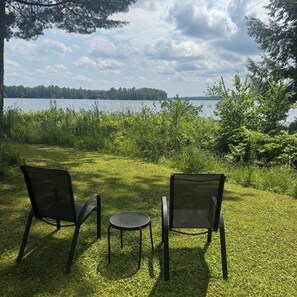 Due sedie e un tavolino in una zona erbosa con vista su un lago e alberi.