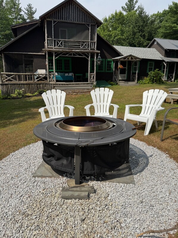Un foyer circulaire entouré de quatre chaises Adirondack blanches, installé sur un sol de gravier, avec un bâtiment en bois en arrière-plan.