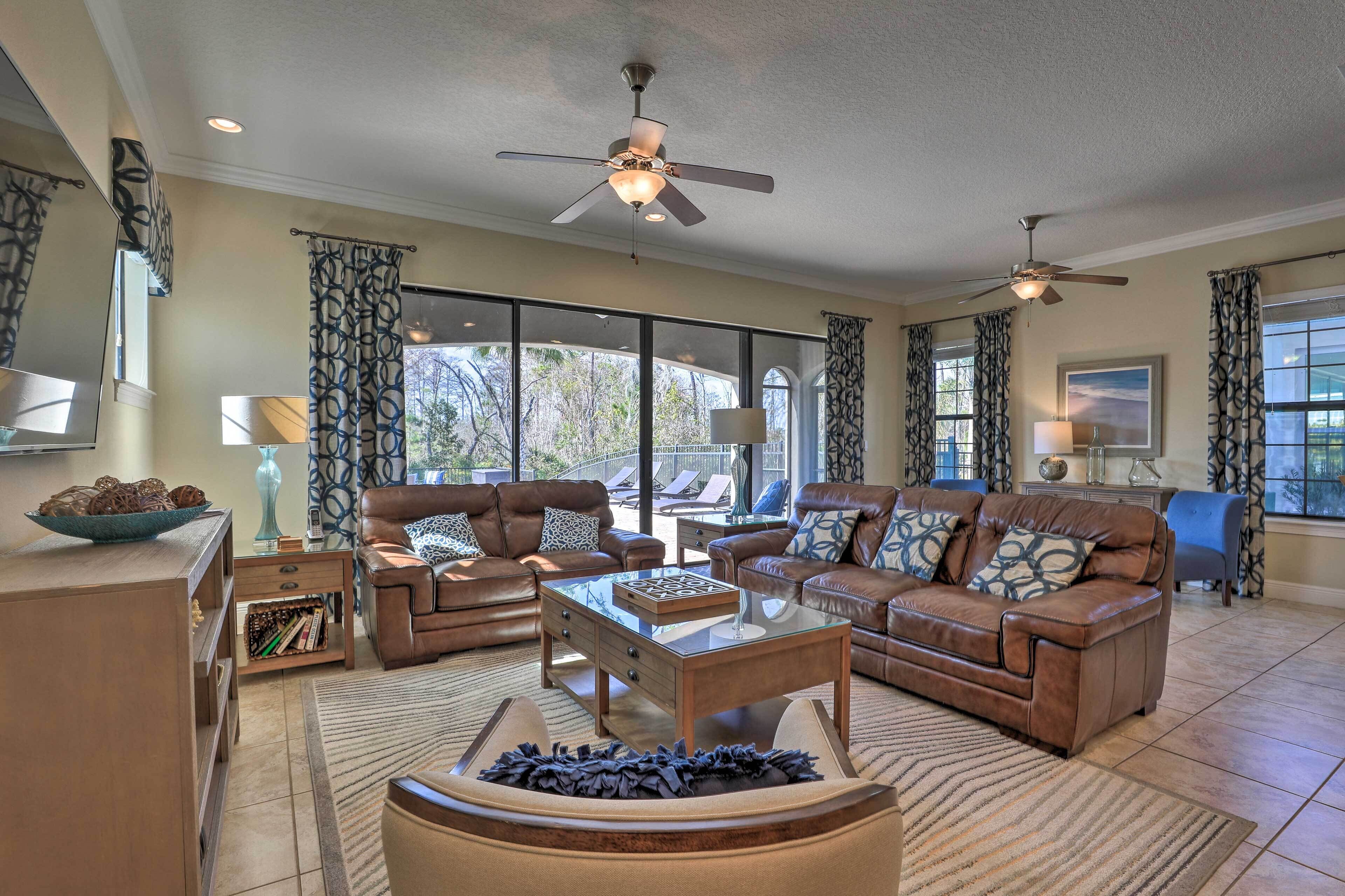 A spacious living room with leather sofas, a glass coffee table, and a ceiling fan. Large windows provide ample natural light and views of the outdoors.