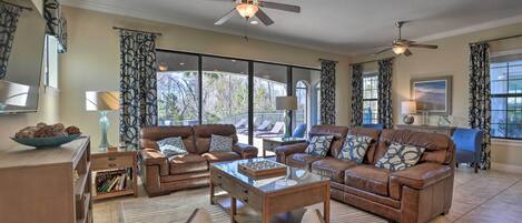 A spacious living room with leather sofas, a glass coffee table, and a ceiling fan. Large windows provide ample natural light and views of the outdoors.