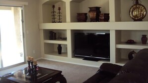 A living room with a flat-screen TV, shelves with decorative items, and a brown leather couch. - *SUMMER SPECIALS** Scenic Golf Course 3 Bed Home w/ Private Pool and Hot Tub (Queen Creek)