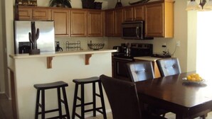 A kitchen with wooden cabinets, a refrigerator, a microwave, a stove, and a dining area with a table and chairs. - *SUMMER SPECIALS** Scenic Golf Course 3 Bed Home w/ Private Pool and Hot Tub (Queen Creek)