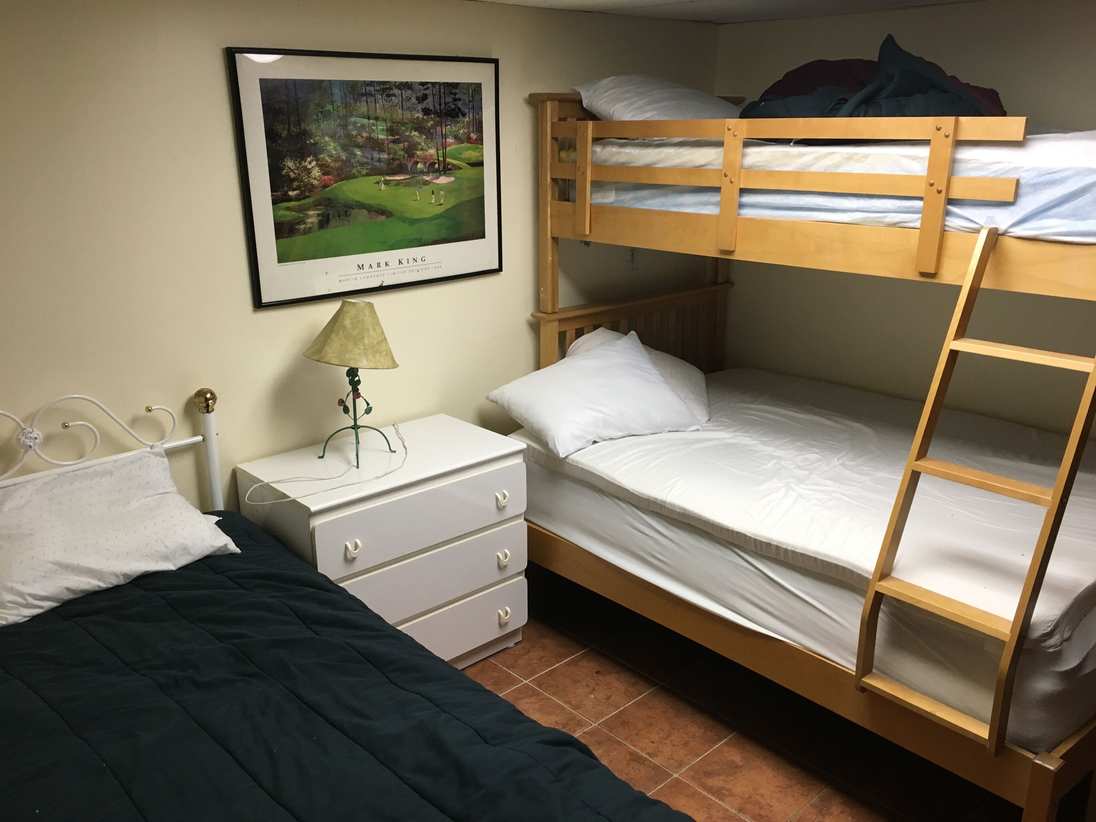 Une chambre avec un lit superposé, un tableau encadré au mur, une table de chevet avec une lampe et une commode.