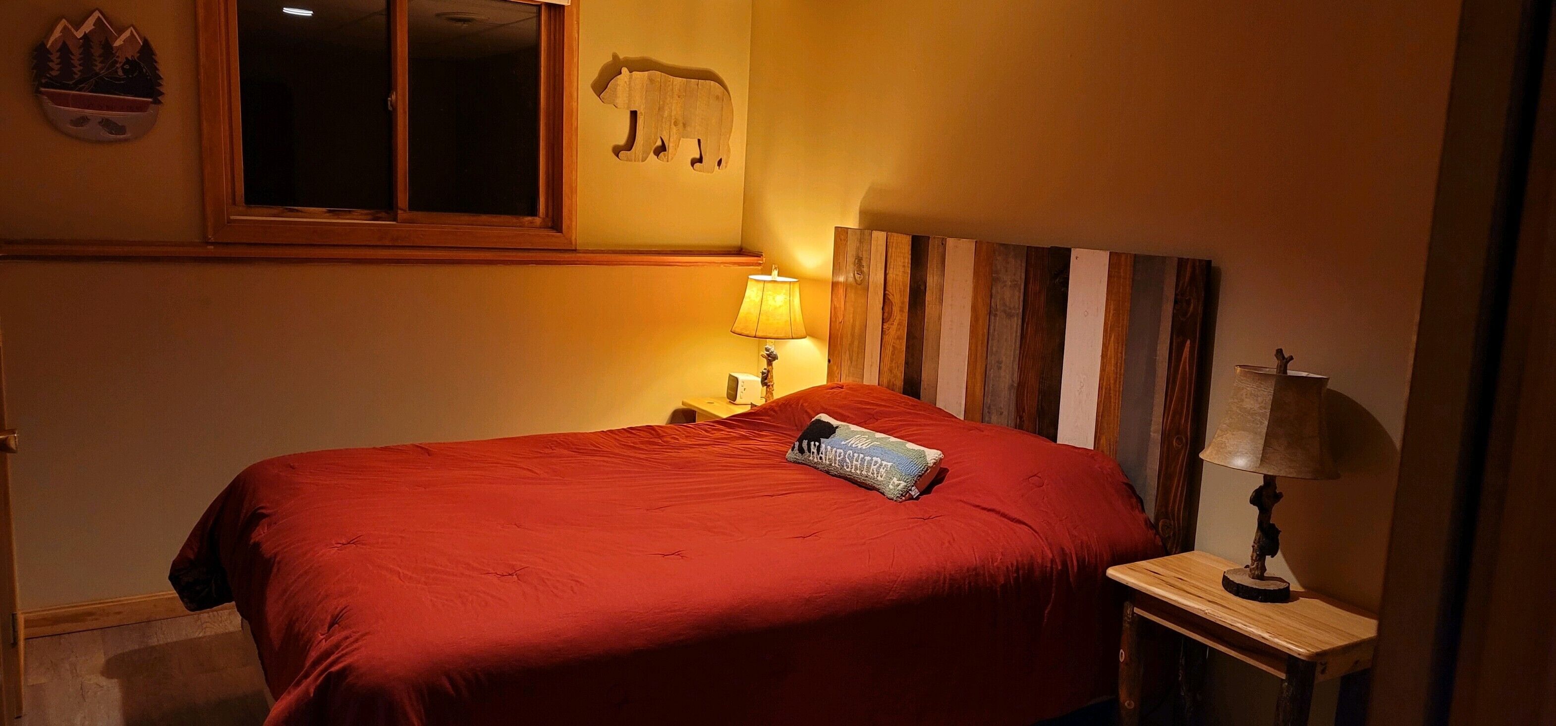 Une chambre à coucher avec une couvre-lit rouge, une tête de lit en bois, une table de chevet avec une lampe et une décoration murale représentant un ours.