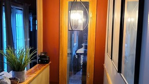Un couloir étroit doté d’un plancher en bois, d’un banc en bois sur lequel est posée une plante en pot, d’un luminaire suspendu et d’une porte donnant sur une autre pièce.