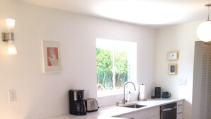 Eine moderne Küche mit einer großen Kochinsel, weißen Schränken, einer Kaffeemaschine und einem Spülbecken. Zwei weiße Stühle stehen an der Insel, und ein Fenster bietet Blick ins Grüne.