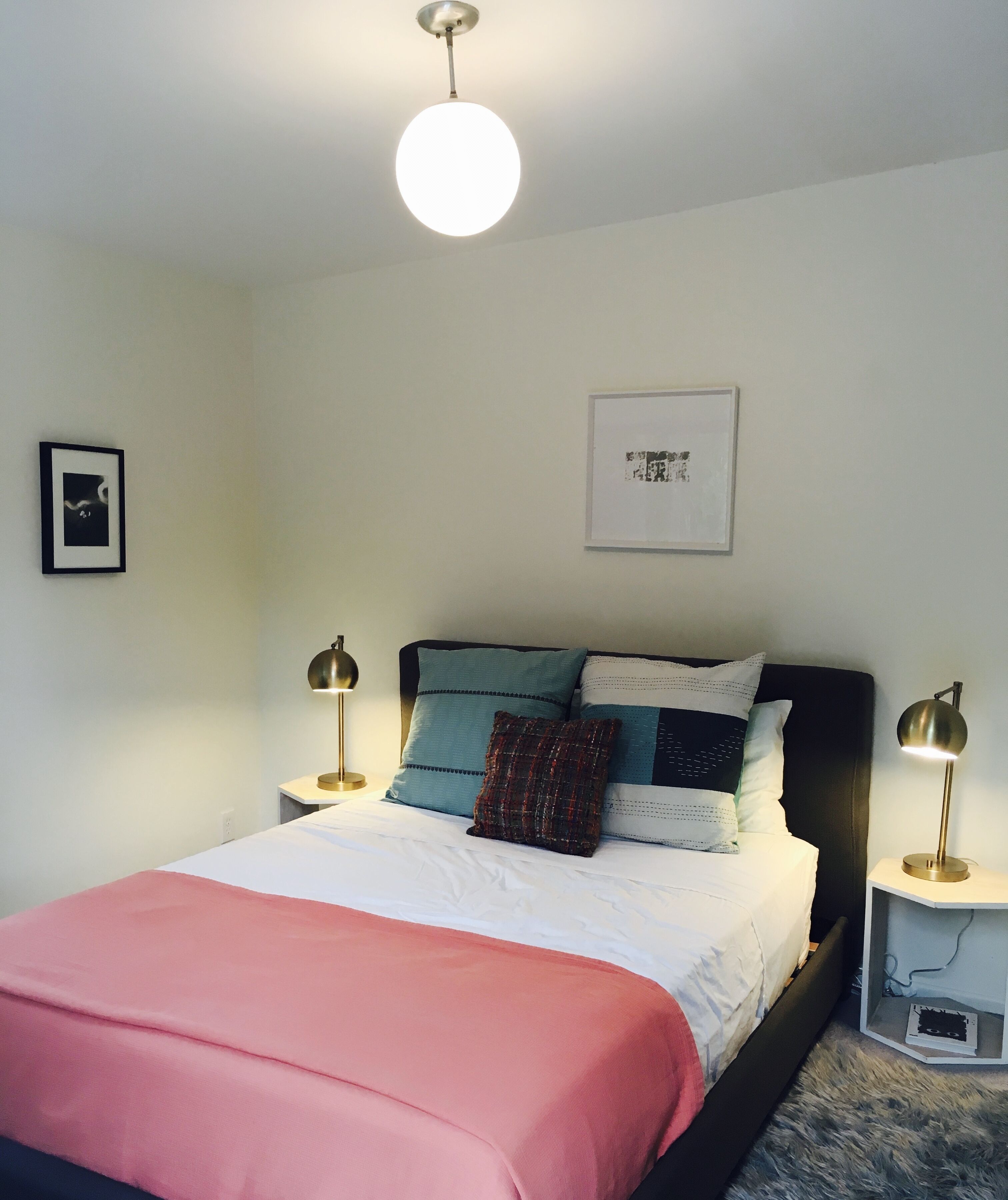 Een slaapkamer met een bed, kussens, een nachtkastje, een lamp en een plafondlamp.