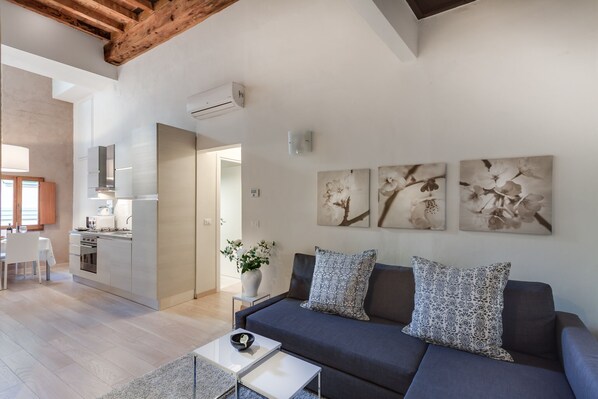 Een moderne woonkamer met een donkere bank, een witte salontafel en een vaas met bloemen. In de achtergrond een keuken met witte kastjes en een eethoek met een witte tafel en stoelen.