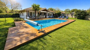 Ein rechteckiger Pool mit einer Holzterrasse, umgeben von einem gepflegten Rasen und einem Haus mit einem Ziegeldach.