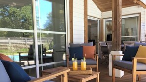Ein überdachter Außenbereich mit Holzmöbeln, einem Tisch mit Getränken und großen Fenstern, die auf einen Garten blicken.