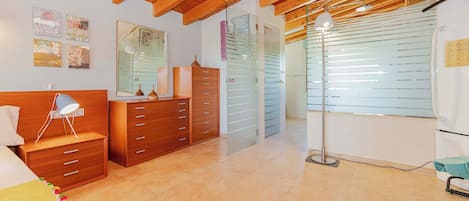 Quarto com teto de madeira, uma cama com travesseiro branco, mesa de cabeceira, cômoda e espelho.