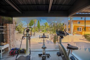 A gym with stationary bikes, a TV, and a view of a courtyard with trees and buildings. - Exclusiva Villa con piscina climatizada, jacuzzi, sauna, gim, y 10000m2 jardín (Alcudia)