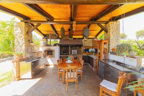 An outdoor kitchen with a stone floor, wooden furniture, and a variety of cooking utensils. - Exclusiva Villa con piscina climatizada, jacuzzi, sauna, gim, y 10000m2 jardín (Alcudia)