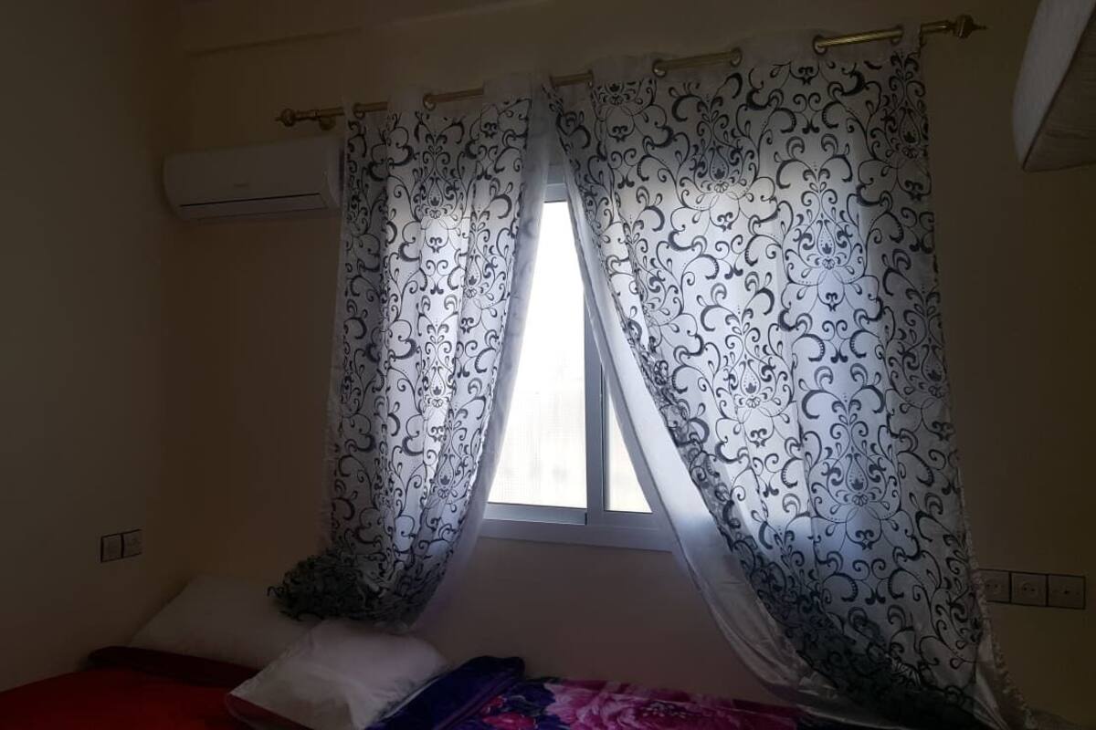 Une chambre avec un lit simple, une fenêtre ornée de rideaux à motifs et un climatiseur fixé au mur.