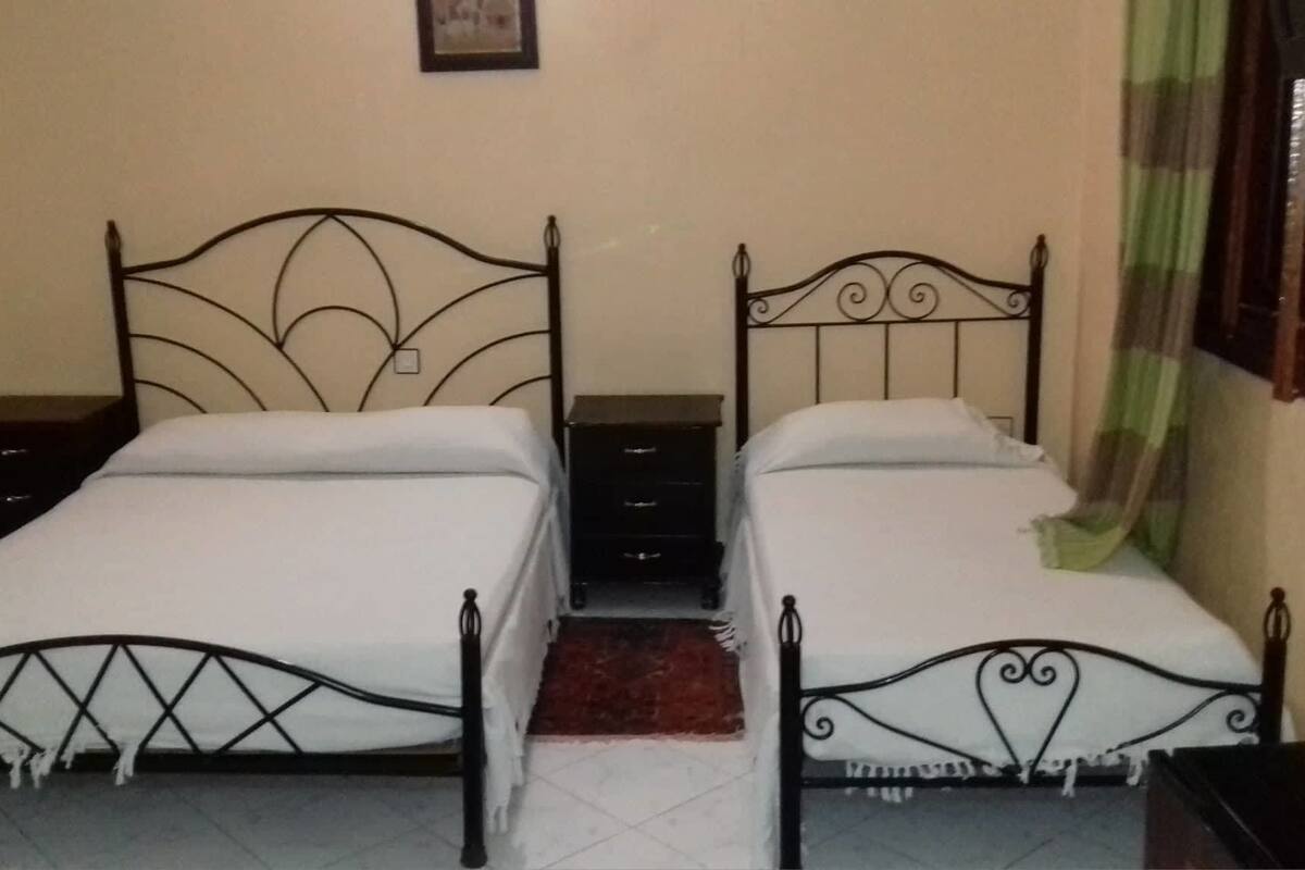 Une chambre avec deux lits, chacun doté d’un cadre en métal et de draps blancs. On y trouve une petite table de chevet en bois, avec des tiroirs situés sous chaque lit.