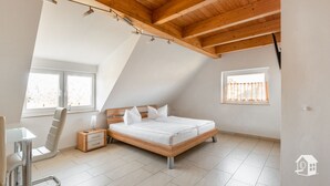 A modern bedroom with a sloped ceiling, a wooden bed frame, a bedside table, a chair, and two windows.