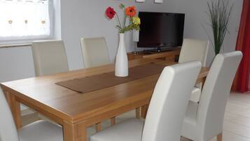 A dining area with a wooden table, white chairs, a vase with flowers, and a television on a stand.