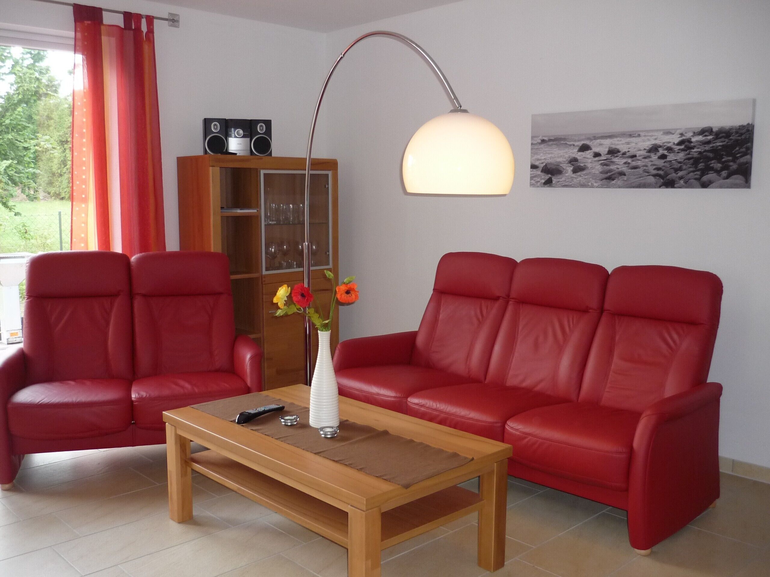 A living room with two red leather sofas, a wooden coffee table, a tall floor lamp, a white vase with flowers, a bookshelf, and a framed picture on the wall.