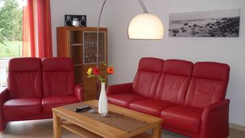 A living room with two red leather sofas, a wooden coffee table, a tall floor lamp, a white vase with flowers, a bookshelf, and a framed picture on the wall.