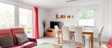 Ein modernes Wohnzimmer mit einem roten Sofa, einem Esstisch mit Stühlen, einem Fernseher auf einem Holzschrank und großen Fenstern mit Vorhängen.