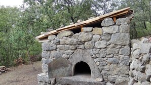 A stone-built outdoor oven with a wooden roof, surrounded by a natural, wooded area.