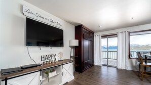 A modern living room with a TV, a console table with plants, a sliding glass door to a balcony, and a sign saying "Lake Times Are The Best Times". - GrandView 305 - Newly Remodeled Waterfront Condo (Chelan)