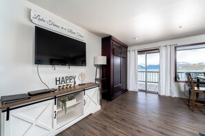 A modern living room with a TV, a console table with plants, a sliding glass door to a balcony, and a sign saying "Lake Times Are The Best Times". - GrandView 305 - Newly Remodeled Waterfront Condo (Chelan)