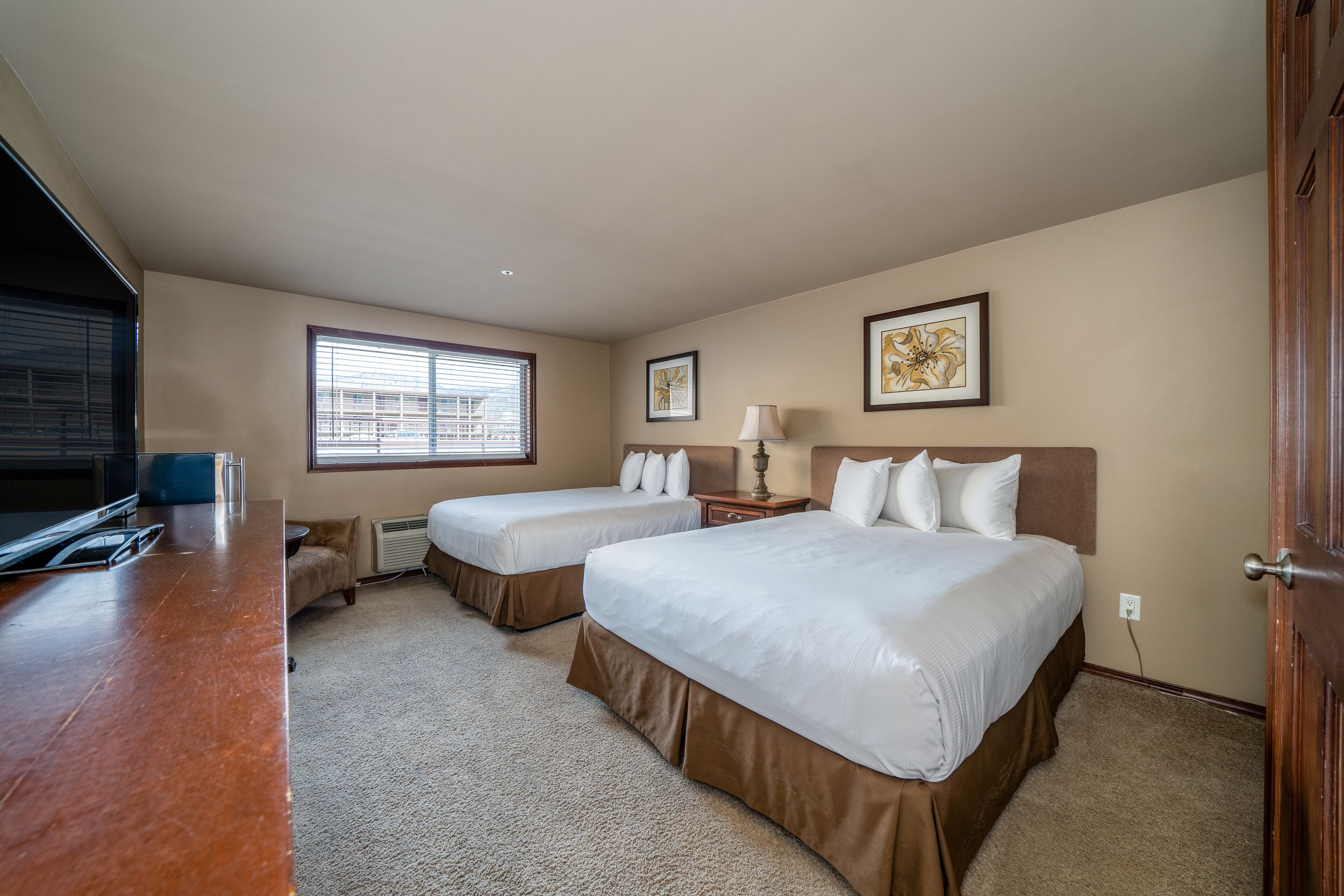 A hotel room with two beds, a desk, a window with blinds, and artwork on the walls.