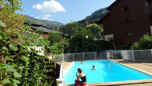 A swimming pool surrounded by greenery, with buildings and mountains in the background. - At the base of the slopes, apartment with balcony/terrace, enclosed garage, nearby shops (Beaufort)