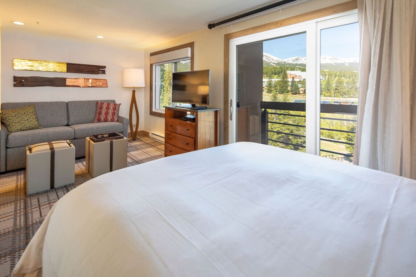 Ein Hotelzimmer mit einem großen Bett, einer Sitzecke, einem Fernseher, einem Schminktisch und einem Balkon mit Bergblick.